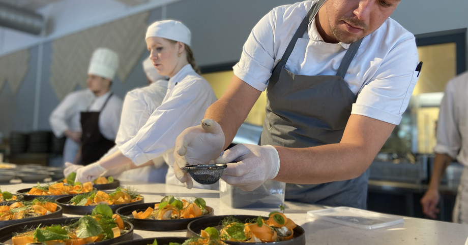 Foto: Hotel- og Restaurantskolen A chef plates food on a dish in a kitchen while other chefs work around him.